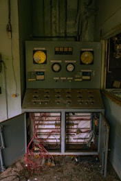 An old, industrial control panel is mounted against a wall, with dials, gauges, and various buttons. Below, tangled wires are exposed inside an open cabinet, suggesting disrepair or neglect. The surrounding area appears dusty and abandoned.