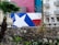 A large mural depicting the Texas flag is painted on the side of a building. The foreground features a stone wall, palm trees, and various other greenery. There are buildings with balconies visible, displaying ornate railings and some bare trees.