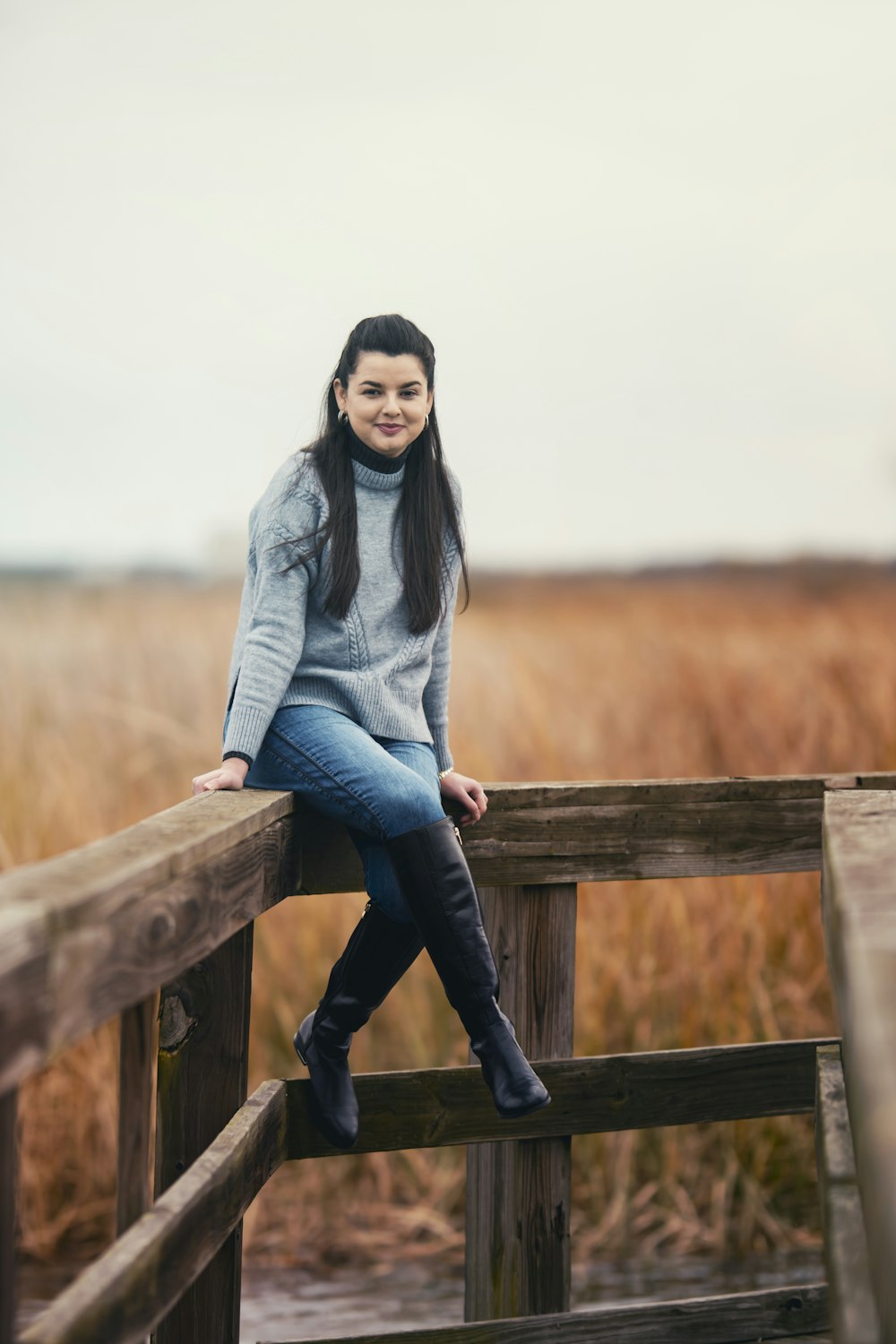 woman in gray sweater and blue denim jeans sitting on brown wooden fence during daytime