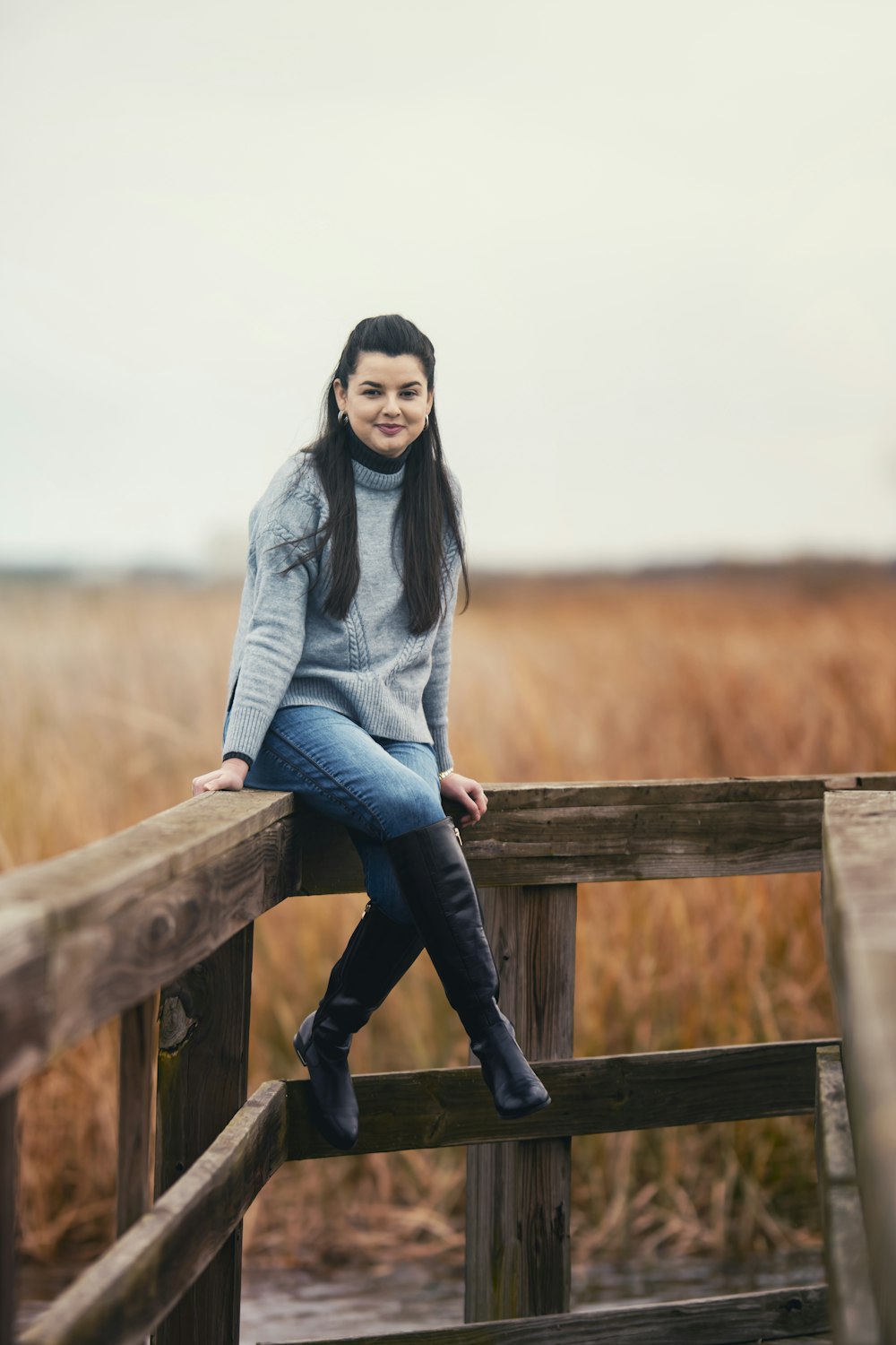 donna in maglione grigio e jeans blu denim seduta su staccionata di legno marrone durante il giorno