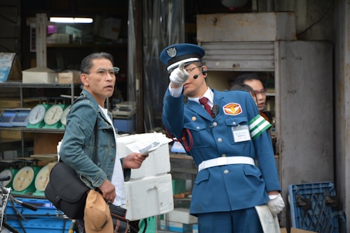 A man in a blue uniform with a cap and white gloves points in a direction while holding a folded document. He appears to be speaking into a headset. Next to him, another man in a denim jacket and glasses holds a cap and a folder, looking in the same direction. Behind them are shelves with weighing scales in an industrial or workshop setting.