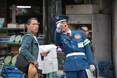A man in a blue uniform with a cap and white gloves points in a direction while holding a folded document. He appears to be speaking into a headset. Next to him, another man in a denim jacket and glasses holds a cap and a folder, looking in the same direction. Behind them are shelves with weighing scales in an industrial or workshop setting.