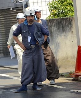 A group of construction workers are standing on a street. They are wearing helmets, vests, and wide pants, with some holding documents. There is a concrete wall and an orange traffic cone nearby.