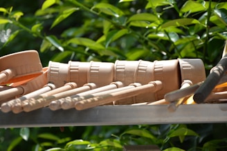 Several beige wooden percussion mallets are arranged in a row, lying on a metallic surface. The background features lush green foliage.