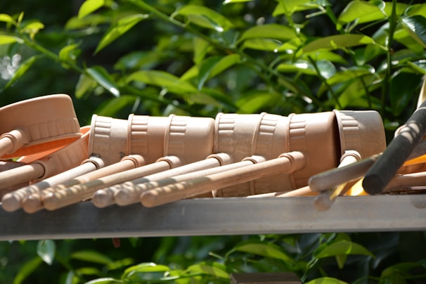 Several beige wooden percussion mallets are arranged in a row, lying on a metallic surface. The background features lush green foliage.
