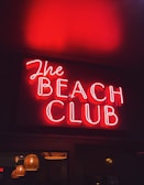 A vibrant red neon sign displaying the words 'The Beach Club', with the glow reflecting warmly on the surrounding area. Below the sign, multiple hanging lamps with a wicker-like texture add a subtle ambient light.