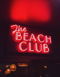 A vibrant red neon sign displaying the words 'The Beach Club', with the glow reflecting warmly on the surrounding area. Below the sign, multiple hanging lamps with a wicker-like texture add a subtle ambient light.