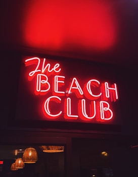 A vibrant red neon sign displaying the words 'The Beach Club', with the glow reflecting warmly on the surrounding area. Below the sign, multiple hanging lamps with a wicker-like texture add a subtle ambient light.