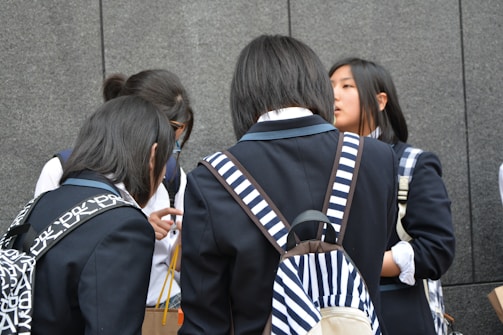 A group of young people wearing school uniforms stands closely together, engaged in conversation. Most of them have backpacks, some with distinctive patterns, and they are positioned against a gray stone wall.