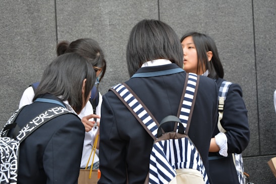 A group of young people wearing school uniforms stands closely together, engaged in conversation. Most of them have backpacks, some with distinctive patterns, and they are positioned against a gray stone wall.