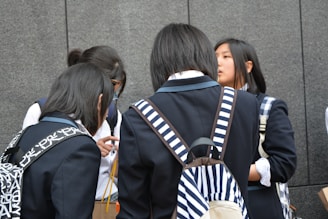 A group of young people wearing school uniforms stands closely together, engaged in conversation. Most of them have backpacks, some with distinctive patterns, and they are positioned against a gray stone wall.