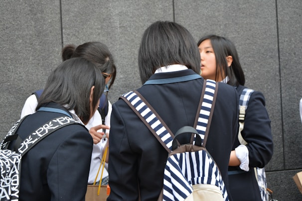 A group of young people wearing school uniforms stands closely together, engaged in conversation. Most of them have backpacks, some with distinctive patterns, and they are positioned against a gray stone wall.