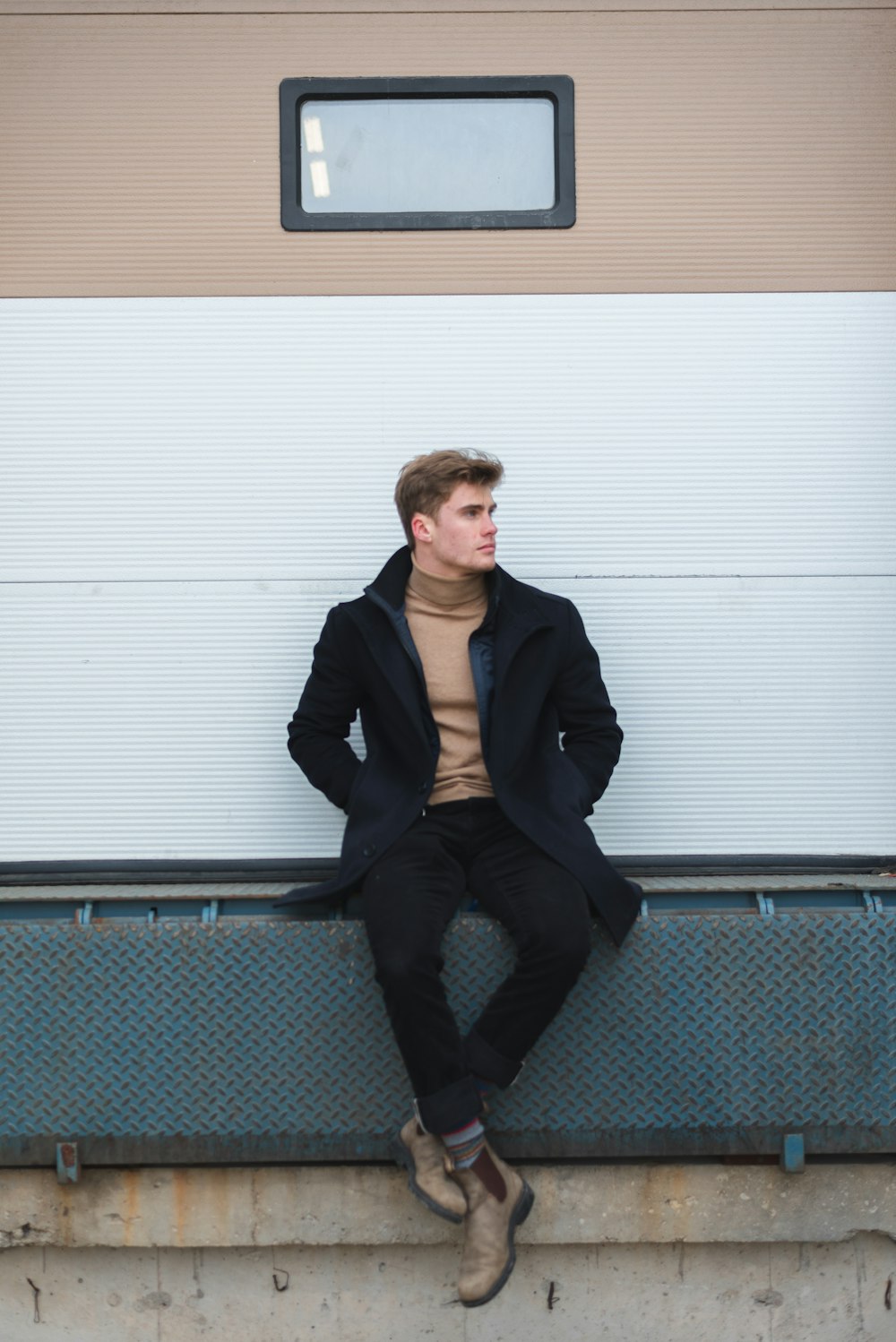 man in black suit jacket and black pants sitting on blue metal fence