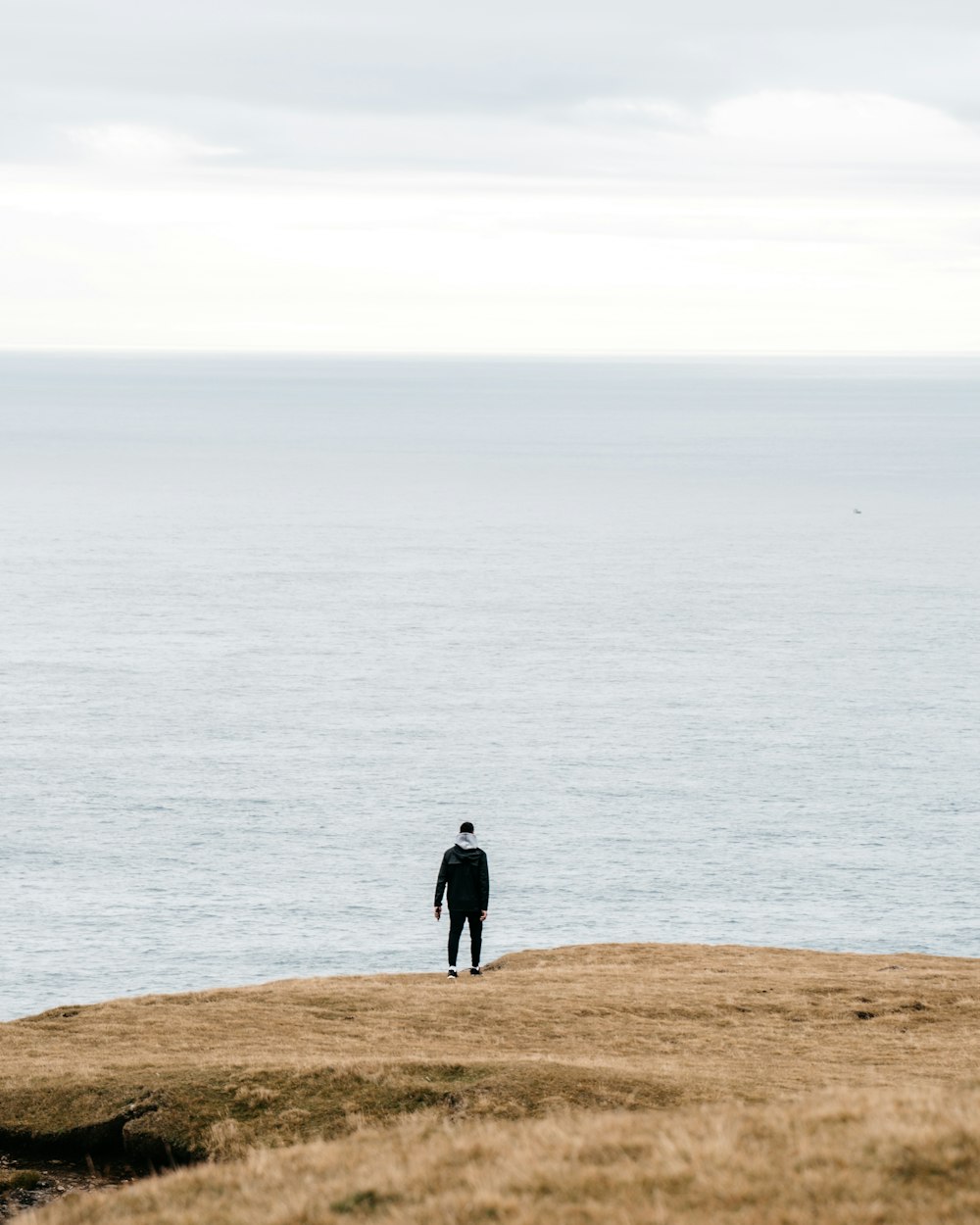 Persona con chaqueta negra de pie en un campo marrón cerca del cuerpo de agua durante el día