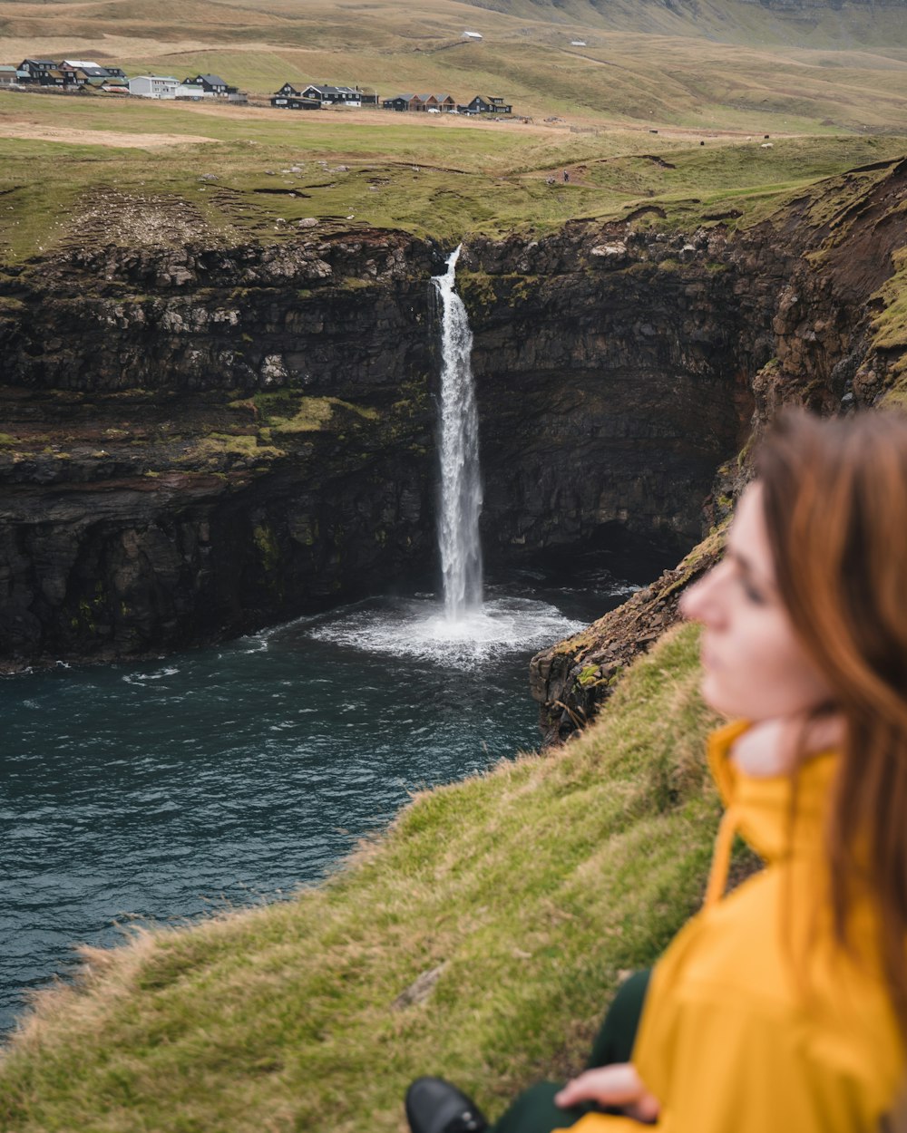 woman in yellow hoodie standing on green grass field near waterfalls during daytime