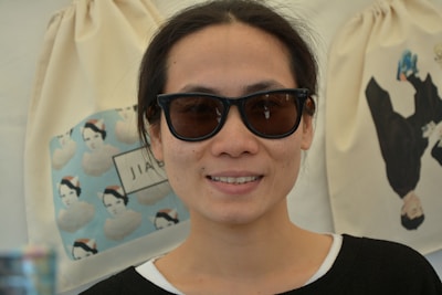 A person wearing dark sunglasses and a black and white top is smiling at the camera. In the background, there are two hanging canvas bags, one of which depicts a pattern of faces with hats and the other shows a person in dark clothing holding a dog.