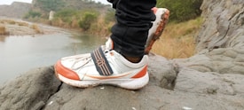A close-up of a person standing on a rocky surface near a stream, wearing orange and white athletic shoes. The background features rugged terrain with sparse vegetation and a small river or stream flowing through the landscape.