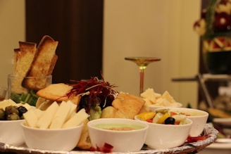 A beautifully arranged charcuterie platter featuring a variety of cheeses, breadsticks, and assorted dips served in small white bowls. Colorful garnishes such as sliced peppers and olives add vibrancy. The setup is elegant and inviting, suitable for a formal gathering.