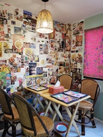 A cozy dining area with a small wooden table and four wicker chairs. The walls are covered with various food magazine clippings and photographs, creating a vibrant collage. A woven pendant light hangs above the table, casting a warm glow. A red tissue box is placed on the table, along with colorful placemats. A window with a pink and patterned curtain is visible to the right.