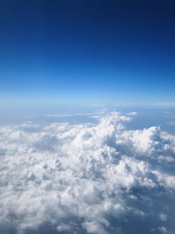 A vast expanse of floating clouds viewed from above, with a clear blue sky extending into the horizon. The clouds appear thick, fluffy, and white, casting soft shadows on the layers below.
