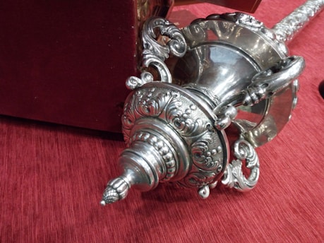 A detailed silver object with intricate ornamental designs lies on a red fabric surface. The object features spiral patterns, curved handles, and a pointed end with elaborate carvings. A portion of another object in a rich dark red color is partially visible in the background.