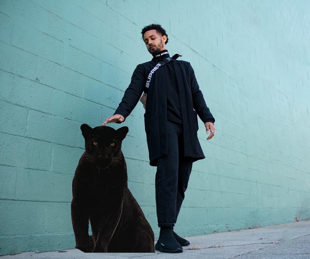 man in black leather jacket and black pants standing beside black short coated dog