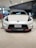 A sleek white sports car is prominently displayed in a modern showroom with polished beige tiles. The front grille and headlight design are eye-catching, and the car has a badge indicating it is a Fairlady Z Nismo model. Surrounding the car are glass windows and additional vehicles.
