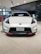 A sleek white sports car is prominently displayed in a modern showroom with polished beige tiles. The front grille and headlight design are eye-catching, and the car has a badge indicating it is a Fairlady Z Nismo model. Surrounding the car are glass windows and additional vehicles.