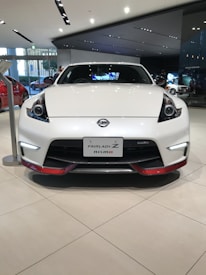 A sleek white sports car is prominently displayed in a modern showroom with polished beige tiles. The front grille and headlight design are eye-catching, and the car has a badge indicating it is a Fairlady Z Nismo model. Surrounding the car are glass windows and additional vehicles.