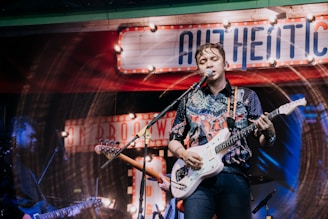 A musician is performing on stage, playing an electric guitar and singing into a microphone. The background is decorated with vintage-style signs and colorful lighting, giving the scene a lively and retro atmosphere. The performer is wearing a patterned shirt and appears to be deeply engaged with the music.
