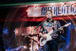 A musician is performing on stage, playing an electric guitar and singing into a microphone. The background is decorated with vintage-style signs and colorful lighting, giving the scene a lively and retro atmosphere. The performer is wearing a patterned shirt and appears to be deeply engaged with the music.