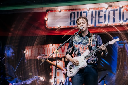 A musician is performing on stage, playing an electric guitar and singing into a microphone. The background is decorated with vintage-style signs and colorful lighting, giving the scene a lively and retro atmosphere. The performer is wearing a patterned shirt and appears to be deeply engaged with the music.
