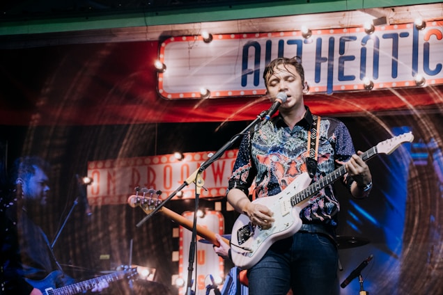 A musician is performing on stage, playing an electric guitar and singing into a microphone. The background is decorated with vintage-style signs and colorful lighting, giving the scene a lively and retro atmosphere. The performer is wearing a patterned shirt and appears to be deeply engaged with the music.