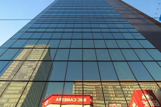 A skyscraper with large glass windows reflects the image of another tall building. The glass panels are arranged in a grid pattern, capturing the clear blue sky and sunlight. Below, there are vibrant red and white signs with logos and text in a foreign language.