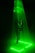 A person stands within a vibrant green laser beam that forms a triangular shape descending from the ceiling to the floor. The beam illuminates the surroundings with a neon glow, casting light patterns on the floor. The person wears a puffy jacket and white sneakers, looking upwards into the light.