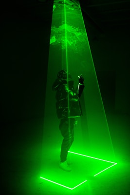 A person stands within a vibrant green laser beam that forms a triangular shape descending from the ceiling to the floor. The beam illuminates the surroundings with a neon glow, casting light patterns on the floor. The person wears a puffy jacket and white sneakers, looking upwards into the light.