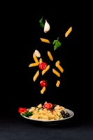 A white plate filled with pasta, cherry tomatoes, olives, and parsley is placed on a black background. Above the plate, pasta, cherry tomatoes, garlic cloves, and parsley leaves are creatively suspended in mid-air, giving a dynamic and artistic presentation of ingredients.
