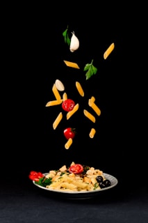 A white plate filled with pasta, cherry tomatoes, olives, and parsley is placed on a black background. Above the plate, pasta, cherry tomatoes, garlic cloves, and parsley leaves are creatively suspended in mid-air, giving a dynamic and artistic presentation of ingredients.