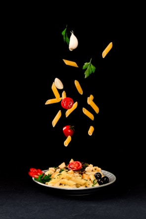 A white plate filled with pasta, cherry tomatoes, olives, and parsley is placed on a black background. Above the plate, pasta, cherry tomatoes, garlic cloves, and parsley leaves are creatively suspended in mid-air, giving a dynamic and artistic presentation of ingredients.