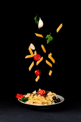 A white plate filled with pasta, cherry tomatoes, olives, and parsley is placed on a black background. Above the plate, pasta, cherry tomatoes, garlic cloves, and parsley leaves are creatively suspended in mid-air, giving a dynamic and artistic presentation of ingredients.