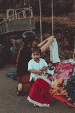 A young child standing near a pile of colorful clothes, holding a red and white dress on a hanger. An adult, possibly a caretaker, wearing a black helmet and dark clothing, is crouched beside them, arranging the clothes. The scene appears to be outdoors, with a metal structure and a blurred natural background.