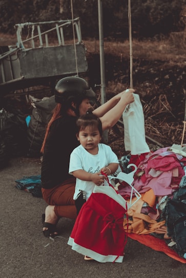 A young child standing near a pile of colorful clothes, holding a red and white dress on a hanger. An adult, possibly a caretaker, wearing a black helmet and dark clothing, is crouched beside them, arranging the clothes. The scene appears to be outdoors, with a metal structure and a blurred natural background.