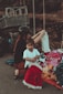 A young child standing near a pile of colorful clothes, holding a red and white dress on a hanger. An adult, possibly a caretaker, wearing a black helmet and dark clothing, is crouched beside them, arranging the clothes. The scene appears to be outdoors, with a metal structure and a blurred natural background.