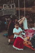 A young child standing near a pile of colorful clothes, holding a red and white dress on a hanger. An adult, possibly a caretaker, wearing a black helmet and dark clothing, is crouched beside them, arranging the clothes. The scene appears to be outdoors, with a metal structure and a blurred natural background.