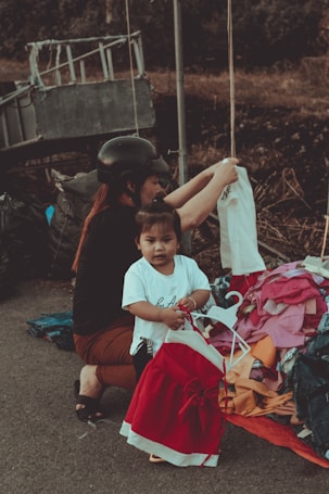 A young child standing near a pile of colorful clothes, holding a red and white dress on a hanger. An adult, possibly a caretaker, wearing a black helmet and dark clothing, is crouched beside them, arranging the clothes. The scene appears to be outdoors, with a metal structure and a blurred natural background.