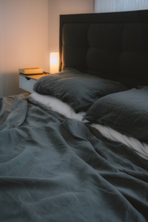 A neatly made bed with dark, soft bedding is placed against a padded headboard. A bedside table holds a small stack of books and a glowing lamp, casting a warm light.