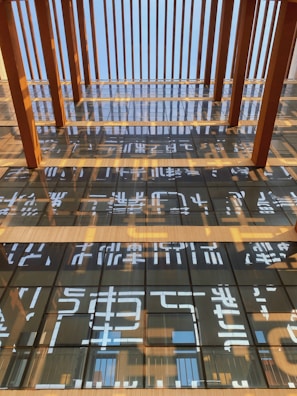 A multi-story glass building facade reflecting a pattern of vertical wooden beams and abstract shapes. The architecture features a grid-like window arrangement with large, reflective panes. Sunlight casts shadows and highlights across the structure, creating dynamic visual textures.