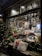 A cozy shop window beautifully decorated for the holidays, with festive lights and intricate designs of snowflakes, Christmas trees, and quaint houses. The interior showcases an assortment of products, including baskets and gourmet items. Outside, two comfortable pillows rest on a wooden bench, surrounded by illuminated Christmas trees.
