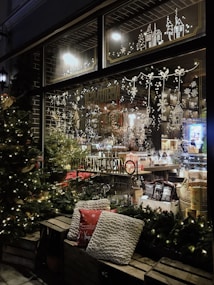 A cozy shop window beautifully decorated for the holidays, with festive lights and intricate designs of snowflakes, Christmas trees, and quaint houses. The interior showcases an assortment of products, including baskets and gourmet items. Outside, two comfortable pillows rest on a wooden bench, surrounded by illuminated Christmas trees.