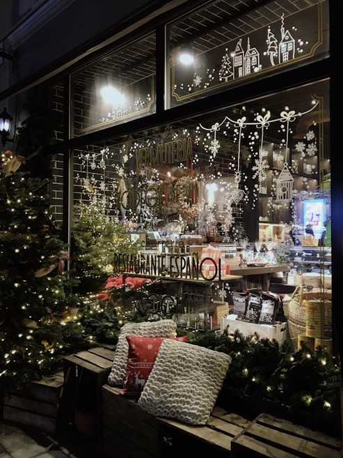 A cozy shop window beautifully decorated for the holidays, with festive lights and intricate designs of snowflakes, Christmas trees, and quaint houses. The interior showcases an assortment of products, including baskets and gourmet items. Outside, two comfortable pillows rest on a wooden bench, surrounded by illuminated Christmas trees.